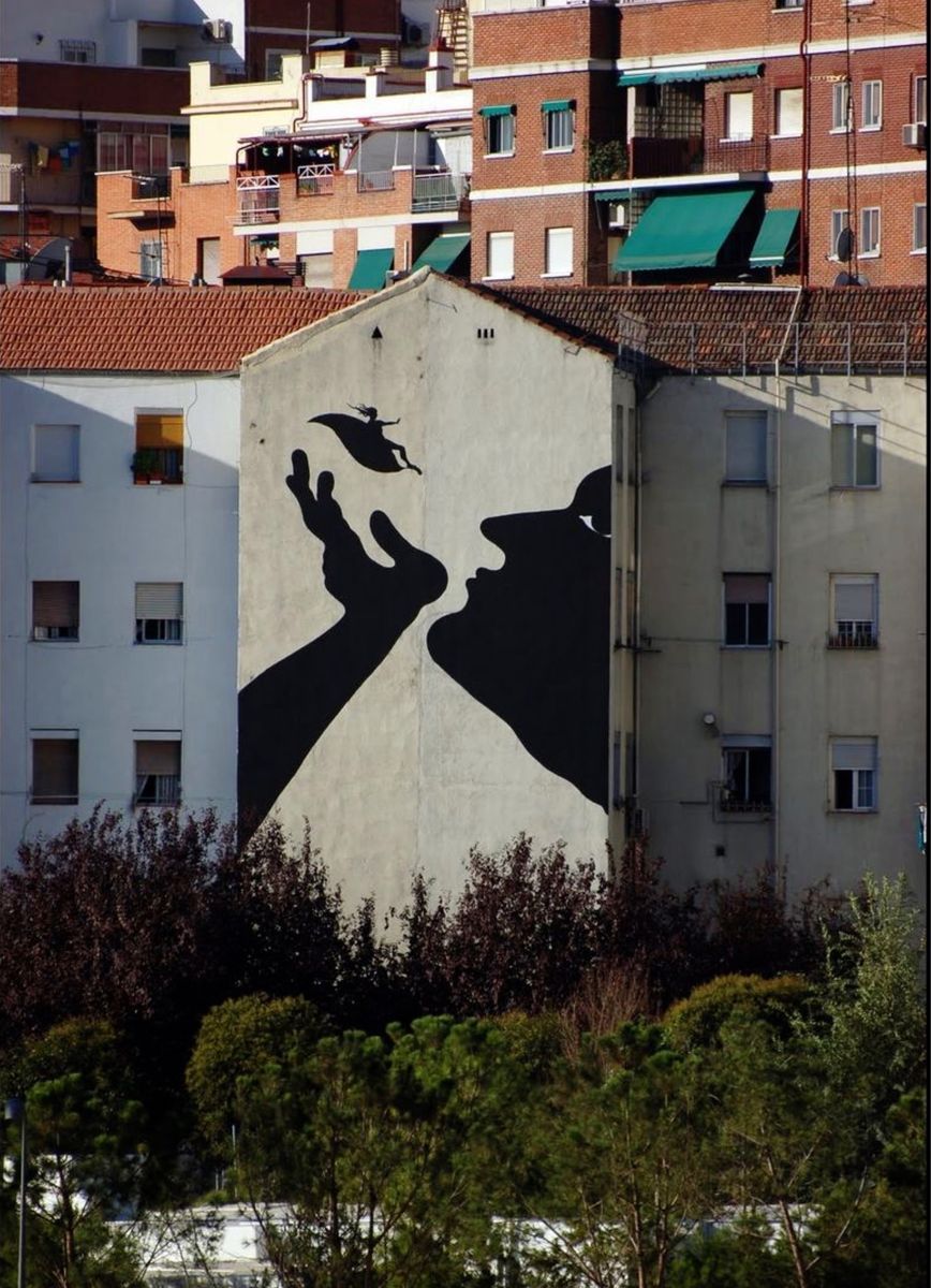 Black silhouette of two enormous profile heads about to kiss, painted across an apartment building gable, a tiny flying fairy figure between their reaching hands, red-brick residential blocks behind.
