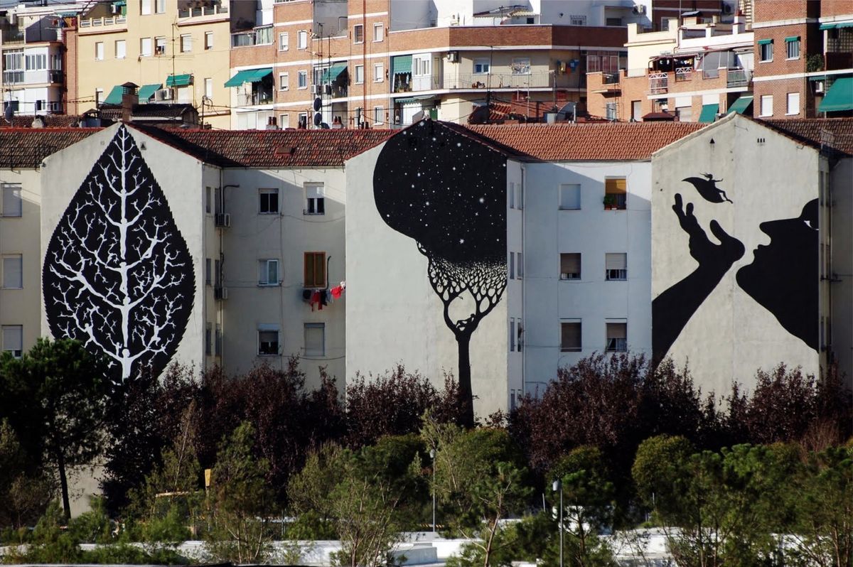 Three adjacent apartment building gables each painted with a different mural: a black leaf with white branching figures, a dark tree silhouette against a starry sky, and two profile heads about to kiss, trees and rooftops in front.