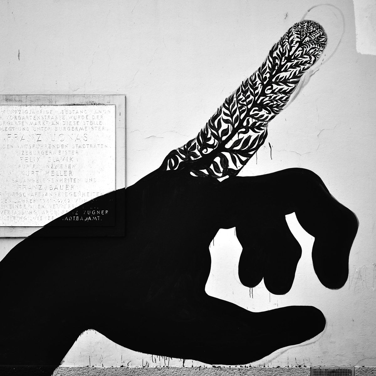 Large black silhouette of a pointing hand, its extended index finger replaced by an intricate black and white leaf pattern, painted beside a commemorative plaque on a plastered wall.