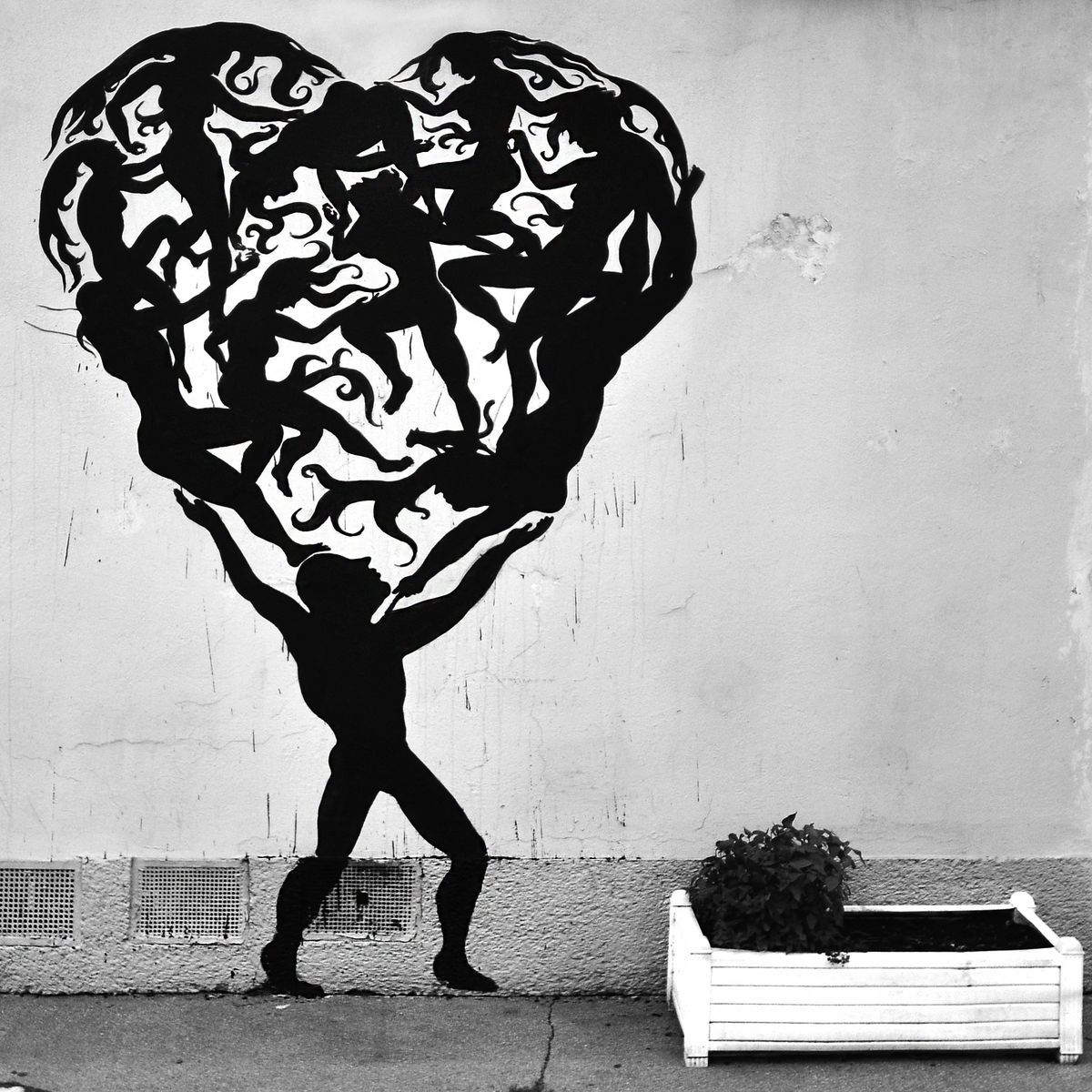 Black silhouette heart made of dozens of intertwined dancing human figures with flame-like patterns, held aloft by a walking figure below, a white planter box on the sidewalk beside it.