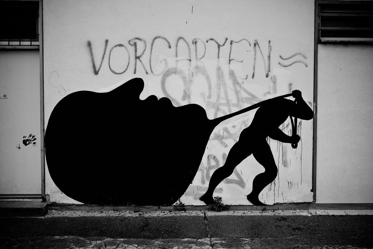 Black silhouette of a bent figure dragging a giant closed eye shape behind it by a rope, the word VORGARTEN spray-painted above on a white wall.