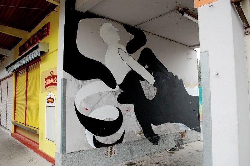 Black and white mural on a building corner showing a reclining nude figure with flowing hair, one leg raised, a swirling ribbon shape curling below, a yellow shop front beside it.