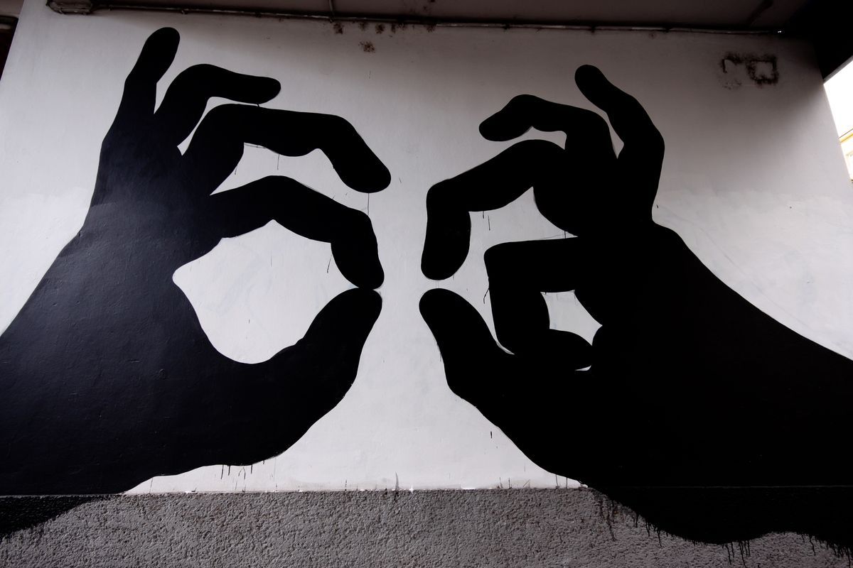 Two large black silhouette hands facing each other, each forming an OK gesture with thumb and forefinger nearly touching, painted on a white wall.