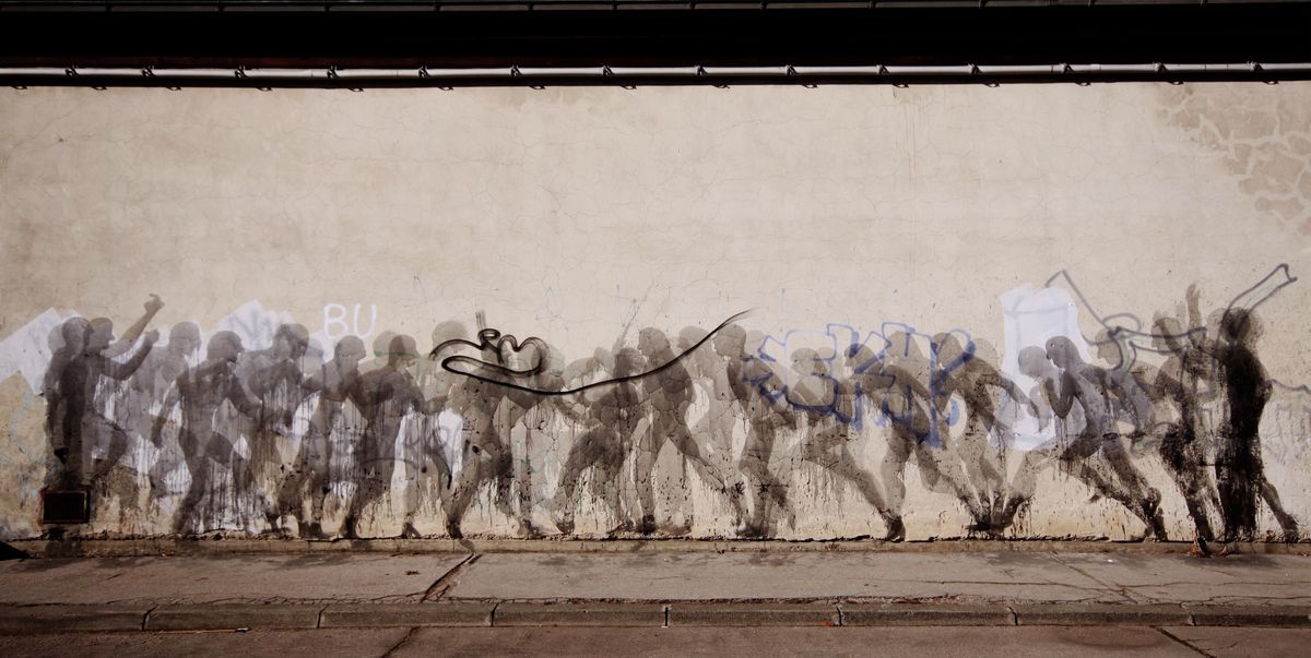 Row of faded gray silhouette figures running in a line across a long plastered wall, graffiti tags and a black scribble mark interspersed among them.