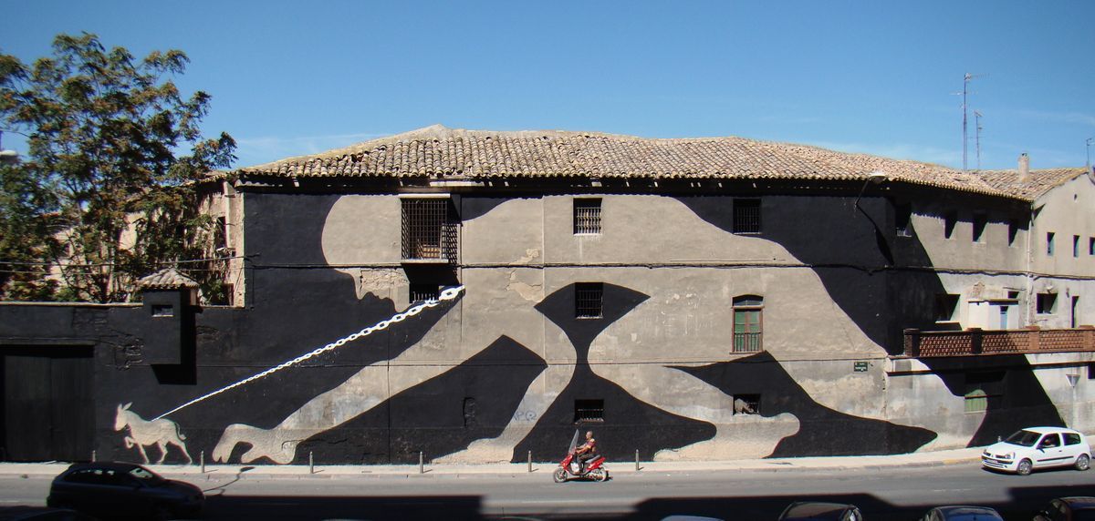 Wide view of the same building corner, the black silhouette figure and hourglass negative space spanning the full facade with several barred windows, a scooter and parked cars passing on the street below.
