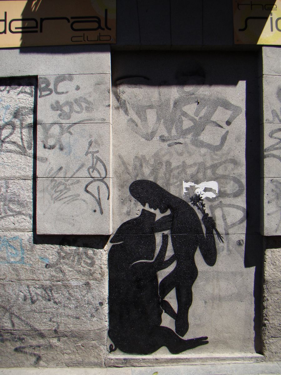 Black silhouette of a seated pig embracing a standing human figure holding a small bouquet, painted on a graffiti-covered wall beneath two shop signs.