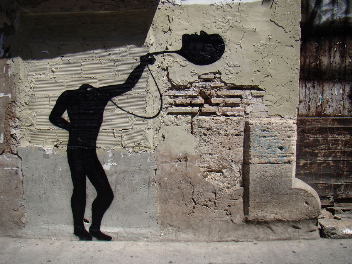 Black silhouette of a bent-over figure with one arm stretched overhead holding a flat oval shape resembling a face or plate, painted on a weathered brick and plaster wall.