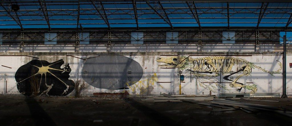 Black splash-shaped mural on a weathered wall resembling an ink blot with radiating lines, silhouetted figures pulling on ropes attached to the lines, one climbing a ladder at the top right.