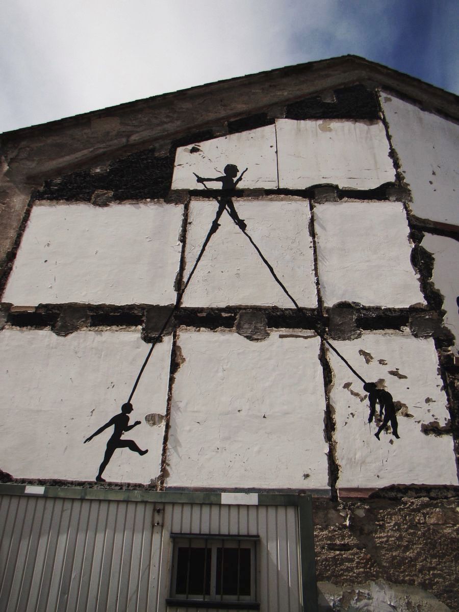 Two silhouetted figures on a gable wall connected by taut ropes to a third figure walking a tightrope near the roofline, one rope figure dangling upside down.