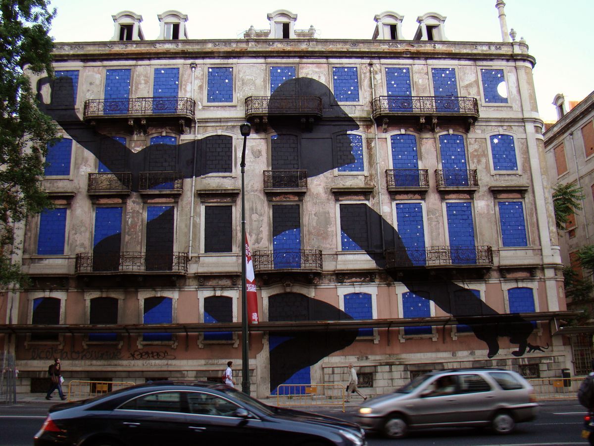Black silhouette of a reclining nude figure sprawled across the blue-tiled facade of an ornate building, its form incorporating a crescent moon and scattered stars painted over the windows, a small cat silhouette at the bottom edge, cars passing on the street below.