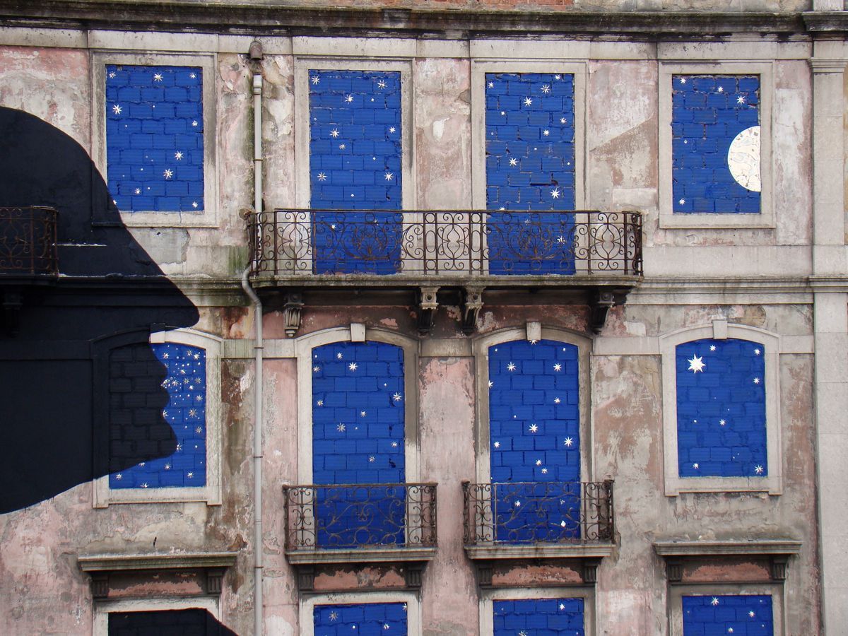 Close-up of the same building facade showing blue-tiled windows painted with white stars and a crescent moon, part of a black silhouette profile head visible at the left edge, wrought iron balcony railings between floors.