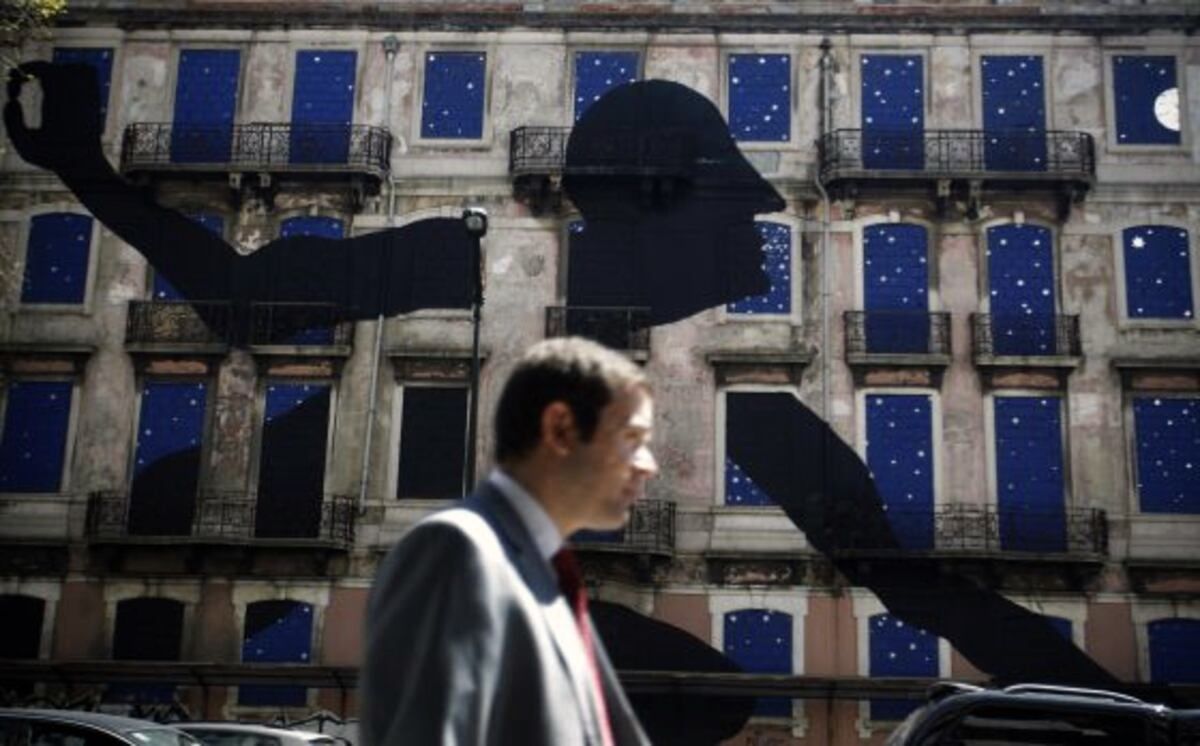 Wide view of the ornate building facade at street level, a black silhouette profile head and outstretched arm stretching across the blue star-painted windows, a man in a suit walking past in front.