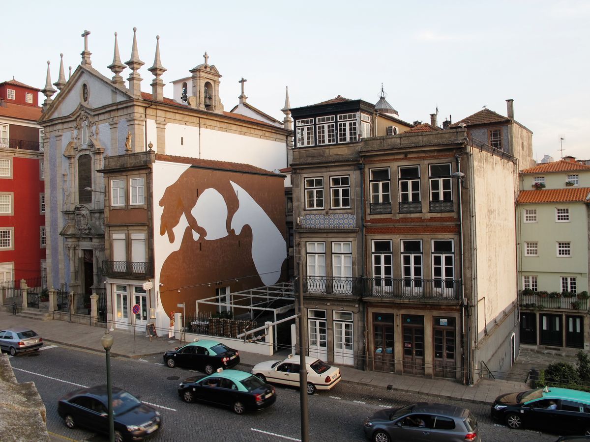Building facade beside a historic church painted with a large brown silhouette of two hands cupped together, seen from above over a street with parked cars.