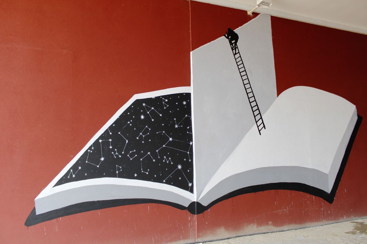 Red wall mural of a giant open book, one page showing a black night sky full of constellations, the other page blank with a black ladder and a small silhouette figure climbing up it.