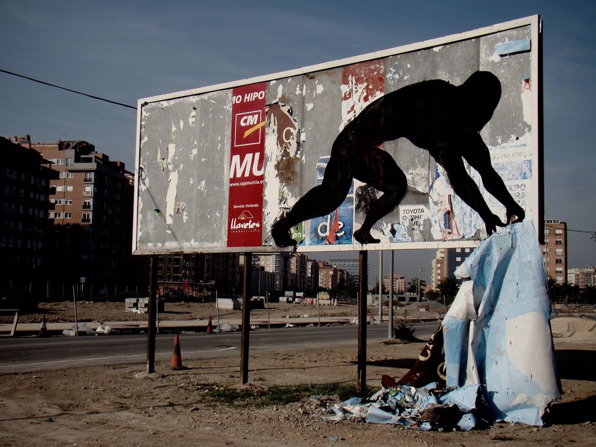 Weathered roadside billboard covered in torn posters, painted with a black silhouette of a crouching figure reaching down toward a crumpled blue tarp on the ground, apartment buildings in the background.