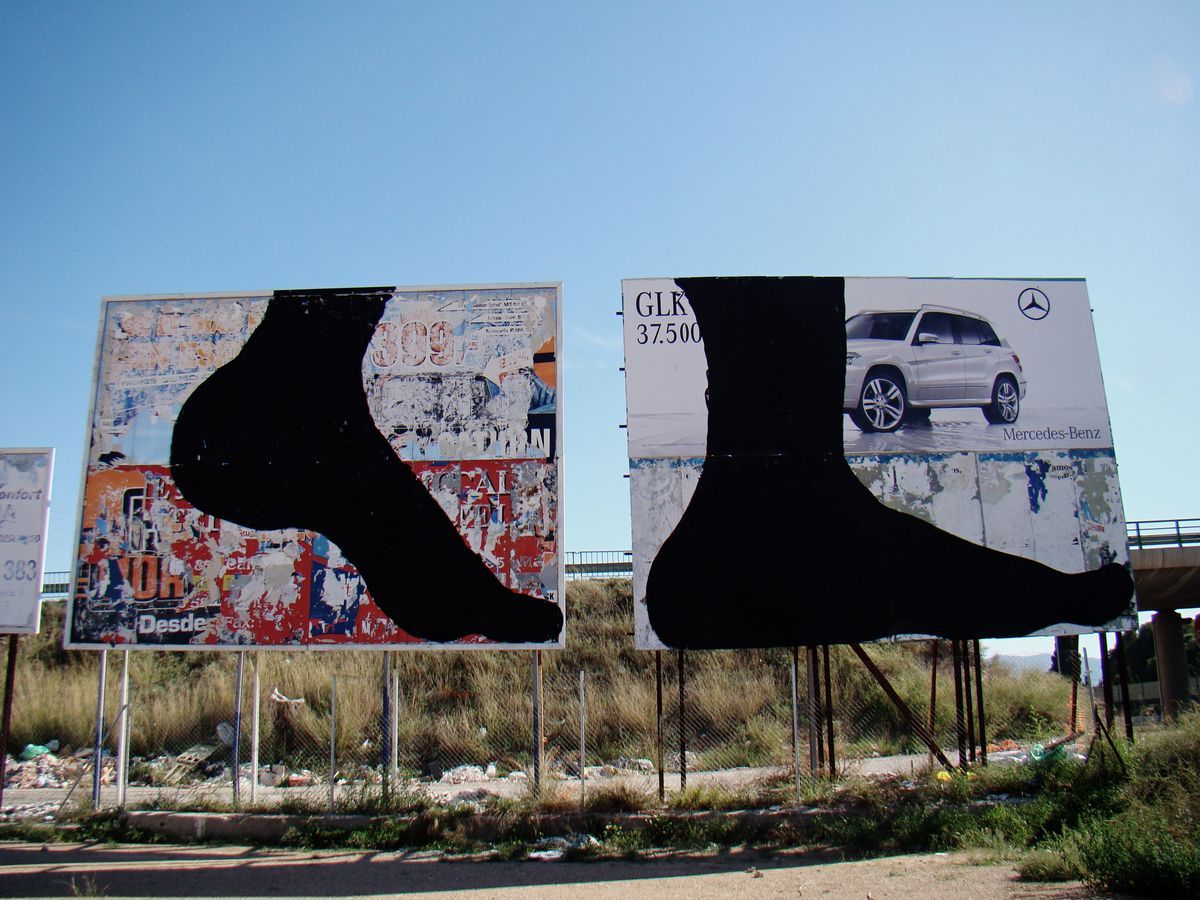 Two billboards side by side, each painted with a large black silhouette of a bare foot stepping down over torn posters, one billboard also showing a Mercedes-Benz advertisement, a highway overpass behind.