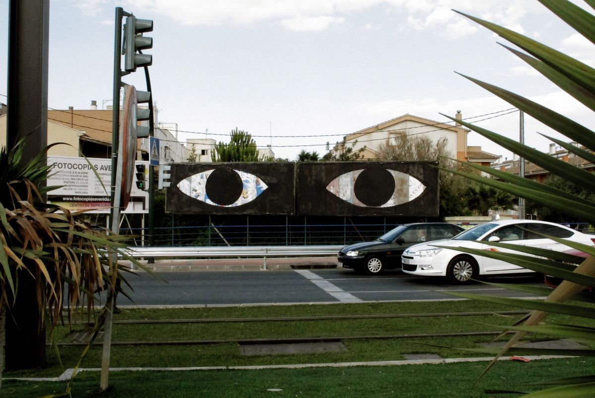 Roadside billboard painted with two large black-and-white eye shapes side by side, traffic lights and parked cars in front, houses and a palm tree in the background.