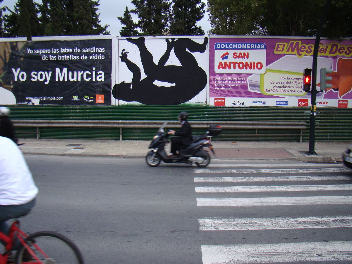 Row of billboards along a street, the center one painted with a black silhouette figure lying on its back with arms and legs raised, a motorcyclist crossing at a pedestrian crossing in front.