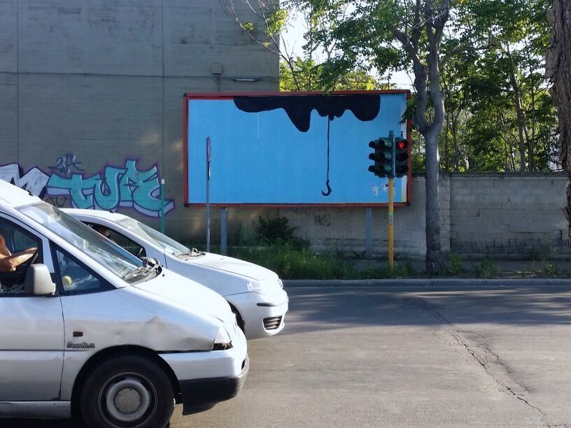 Small light blue billboard painted with a black dripping shape hanging down into a fishing hook, parked cars and trees in front, a traffic light beside it.
