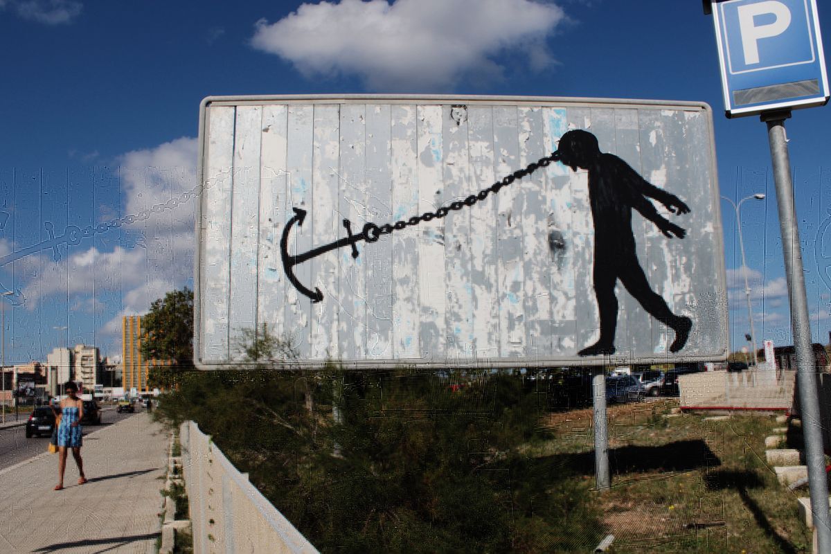 Weathered road sign painted with a black anchor connected by a chain to the mouth of a walking silhouette figure, as if being dragged forward, a parking sign and pedestrian visible on the sidewalk beside it.