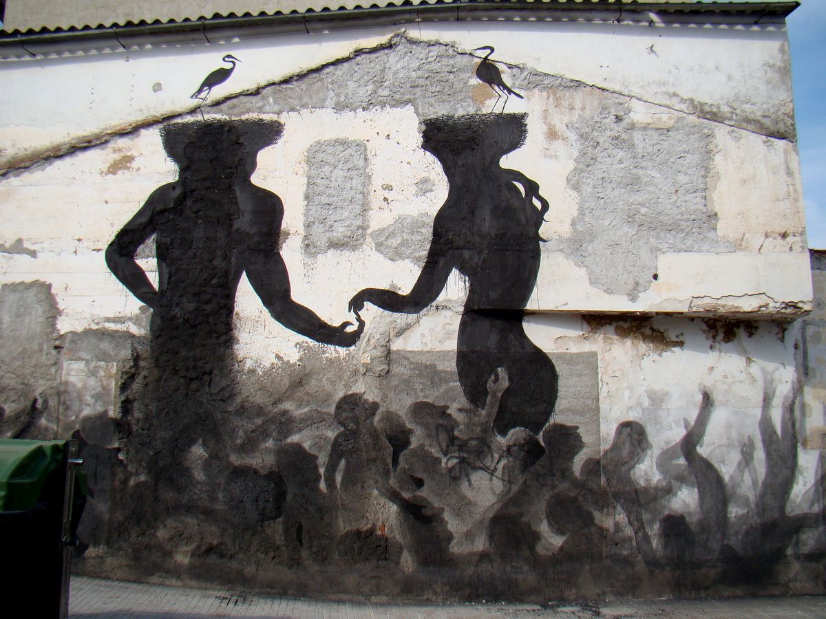 Two black silhouette figures with rough textured heads holding hands on a weathered wall, a heron standing on each head, a crowd of smaller silhouette figures painted below them.