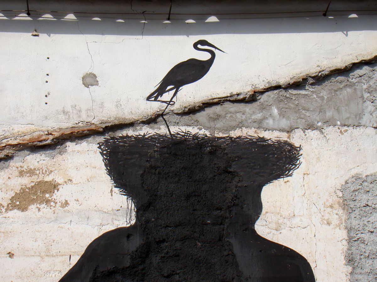 Close-up of one black silhouette figure's rough textured head with a heron standing on top, wings folded, against a cracked plaster wall.