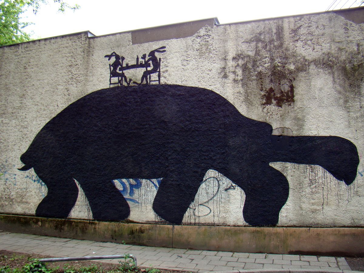 Large black silhouette of a lazy hare-like creature lying across a concrete wall, two small rabbit figures seated at a table on its back, a paved courtyard in front.