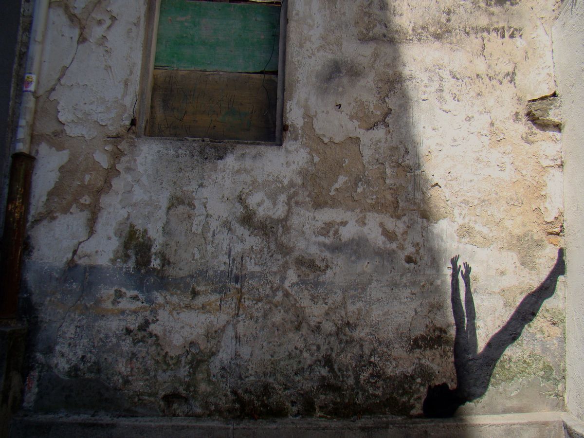 Black silhouette figure with raised hands and a round eclipse-like head, painted on a cracked plastered wall, a white circular pattern on its chest, hand-print graffiti scattered around it.
