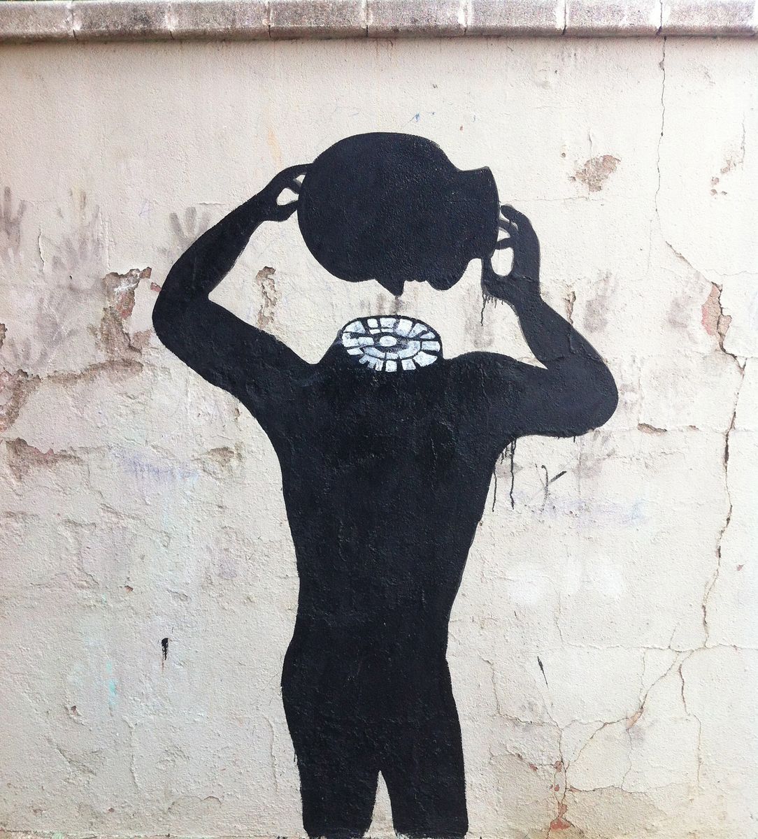 Black silhouette of a walking figure whose head is made of dozens of round balloon-like shapes clustered together, painted on the wall of a ruined roofless building, rubble and a broken window nearby.