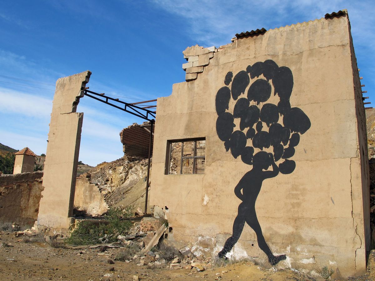 Black silhouette of a person walking with a large cluster of round balloon-like shapes for a head, painted on the wall of a ruined roofless building.