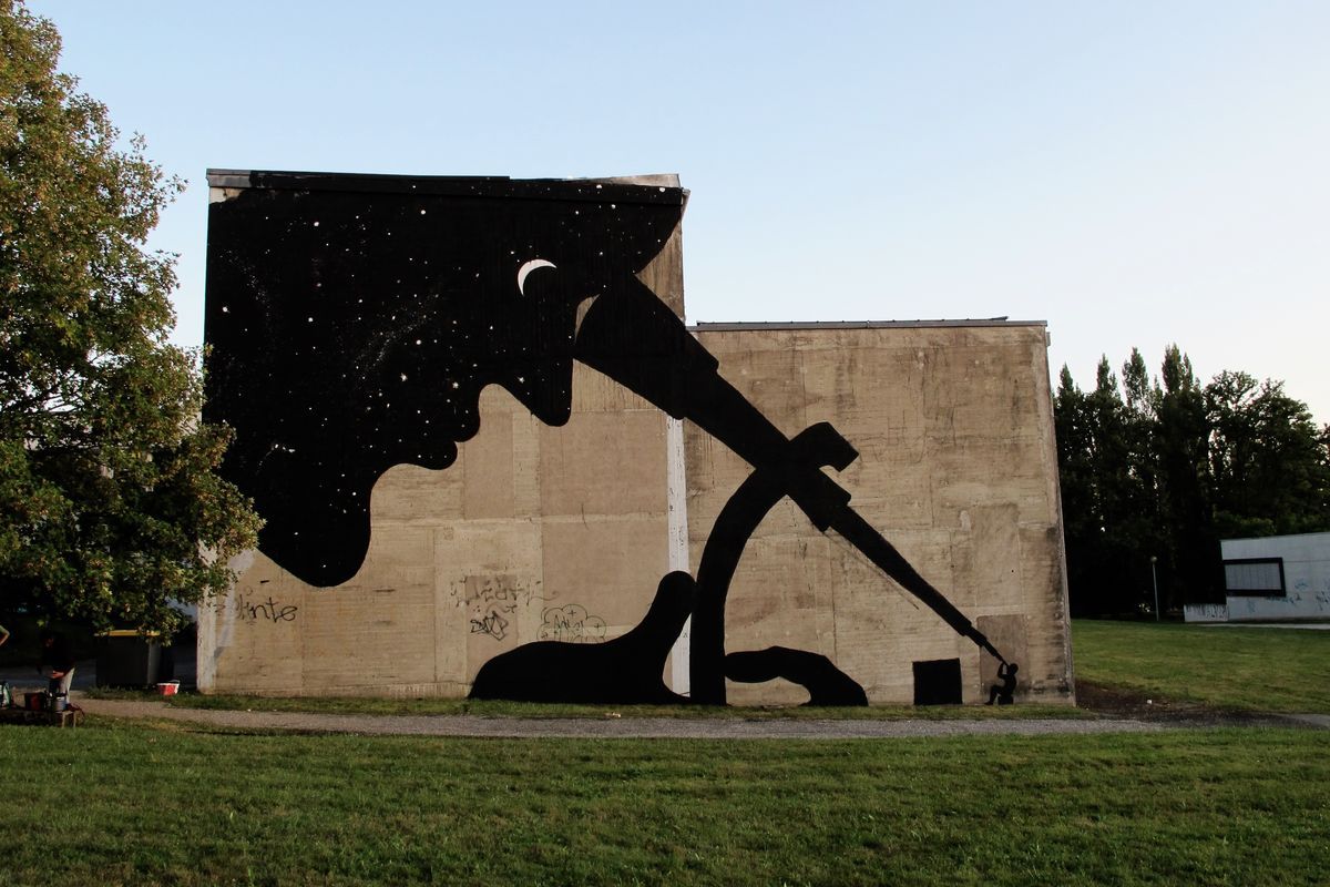 Black mural on a building facade: a silhouette holding a starry sky and a crescent moon stretches into an arm shaped like a telescope, pointing down at a small figure on the ground.