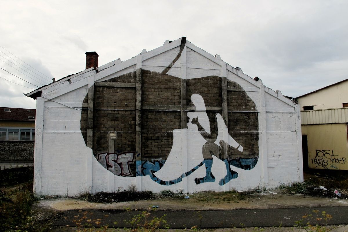 Small gabled white warehouse painted with a large dark oval shape resembling a womb, a white silhouette of a curled fetal figure inside it, graffiti tags along the base of the wall.