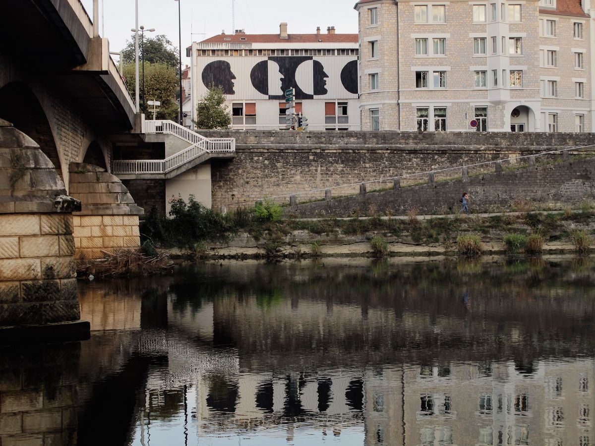 Building facade seen across a river from beneath a stone bridge, painted with four black silhouette profile faces in a row, each overlapping the next like phases of the moon, the mural reflected in the water below.