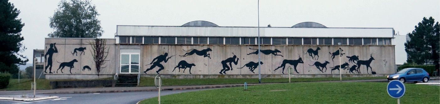 Long low building facade painted with a running black silhouette hunter figure chased by leaping hounds and deer across several panels, two children standing on the grass in front.