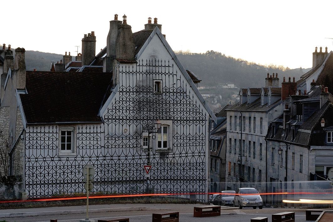 Gabled white house facade entirely covered in a painted trompe-l'oeil pattern resembling ornate black wrought-iron security grilles, a car passing on the street below at dusk.