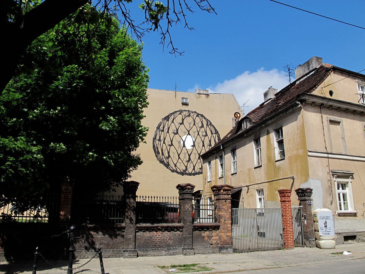 Wide street view of the spherical net mural on a building facade, seen past a large green tree and a residential building with a gated entrance in the foreground.