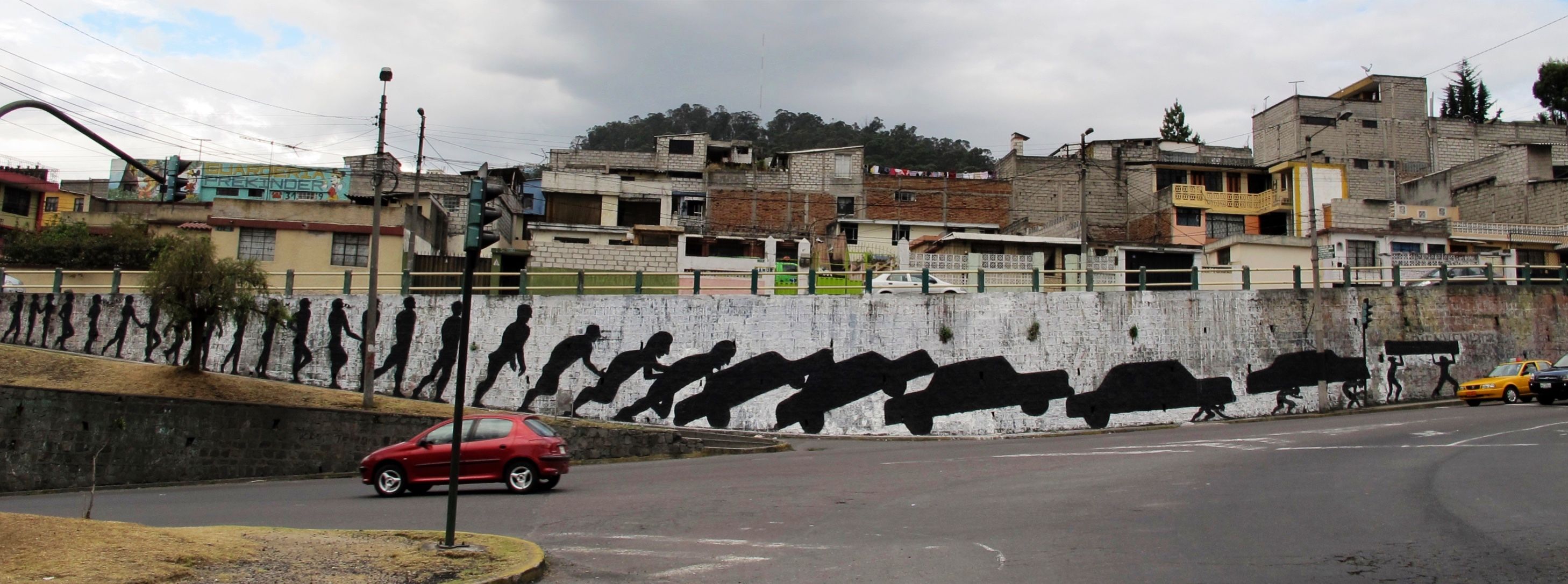 Long retaining wall mural showing a row of walking black silhouette figures gradually transforming into a line of cars, painted along a street corner with houses on the hillside behind, a red car passing by.