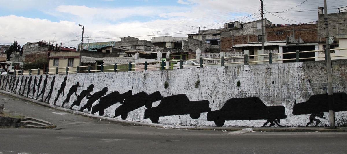 Long retaining wall mural showing a row of walking black silhouette figures gradually transforming into a line of cars, painted along a street corner with houses on the hillside behind, a red car passing by.