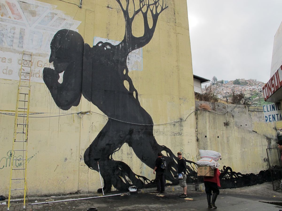 Large black silhouette on a building corner of a bent figure with a tree-like branching form growing from its back, workers and a ladder at the base, hillside houses visible in the distance.
