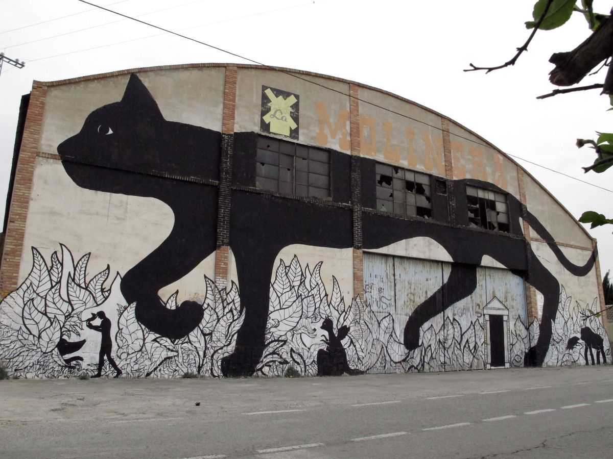 Long warehouse facade painted with a giant black cat walking across broken windows, its tail curling down into a row of black-and-white outlined leaves and small animal silhouettes along the ground.
