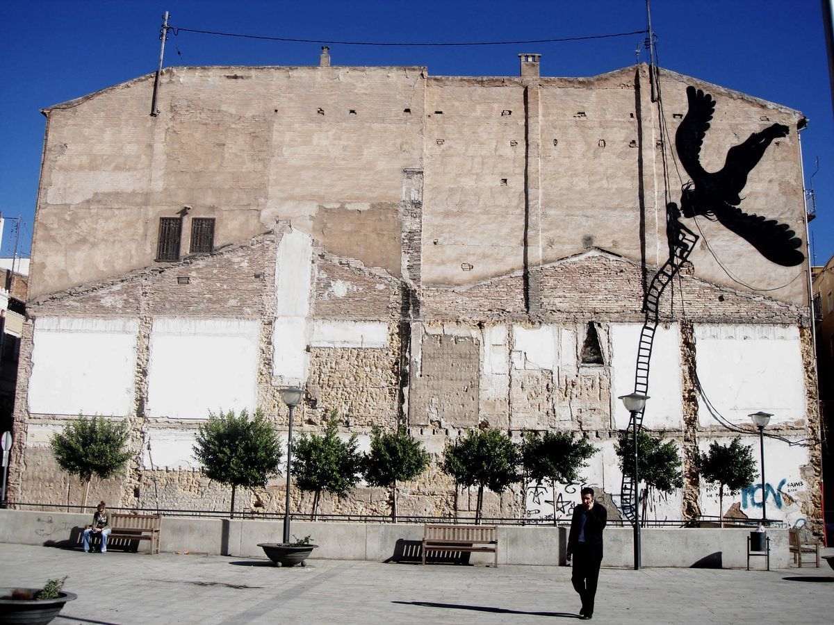Wide view of a ruined building facade with a black silhouette bird in flight near a ladder on the upper right corner, a plaza with benches, young trees, and a man walking below.