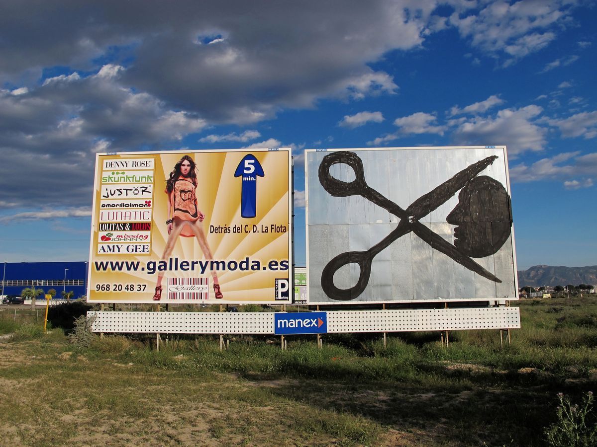 Roadside billboard painted with a black silhouette of a police baton or club shape, an abandoned farmhouse and palm tree beside it, a corrugated metal fence running along the background.