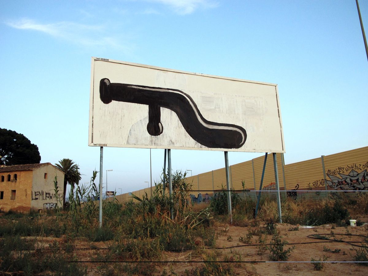 Black silhouette resembling a bent pipe or spigot painted across a large roadside billboard, weeds and an abandoned farmhouse in the foreground, a corrugated metal fence running behind.