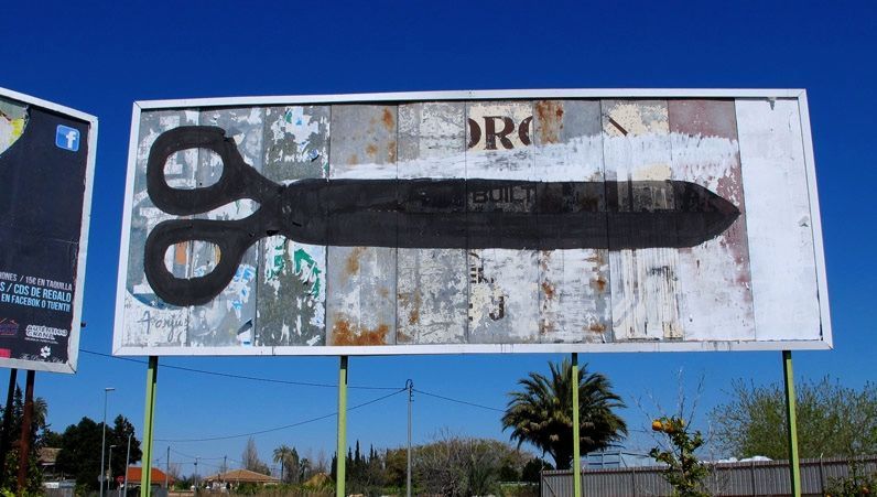 Closer view of the same rusted billboard showing the black scissors alone, blades closed into a single elongated shape pointing right, without the hanging figures.