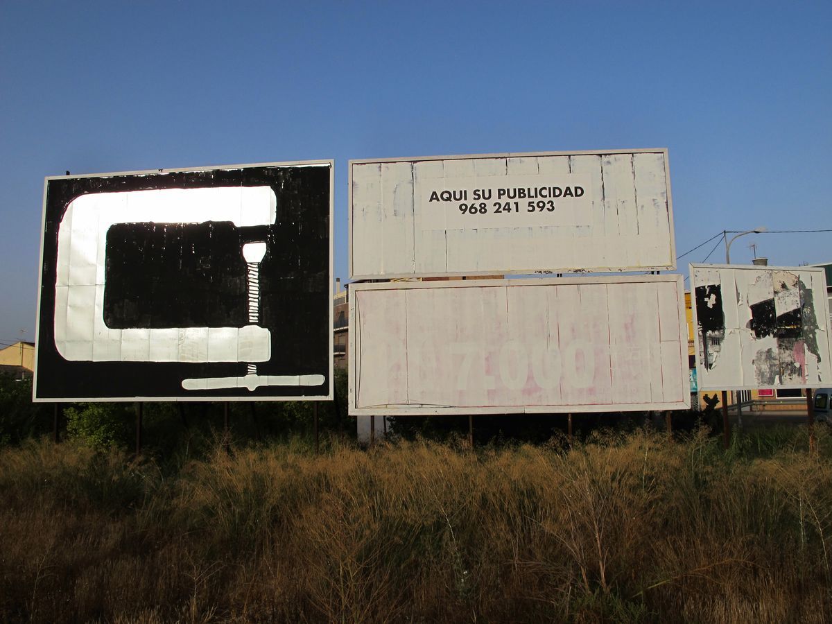 Row of blank billboards beside a road, the nearest one painted black with a white C-shaped cutout and a small vertical line, the others left empty with a placeholder phone number and torn poster fragments.