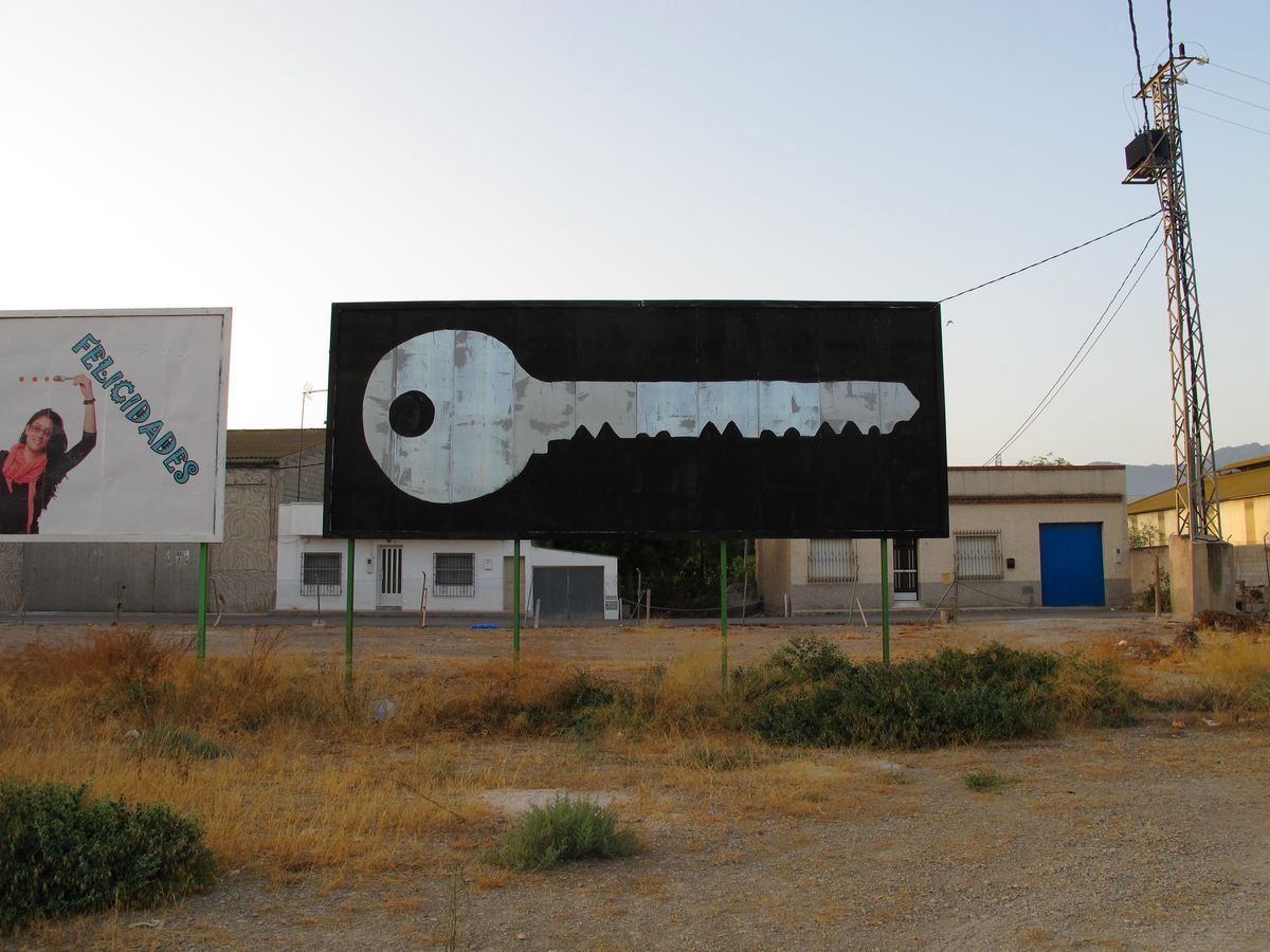 Black and white billboard painted as a large house key with a round head and serrated teeth, standing on green poles beside another sign reading Felicidades.