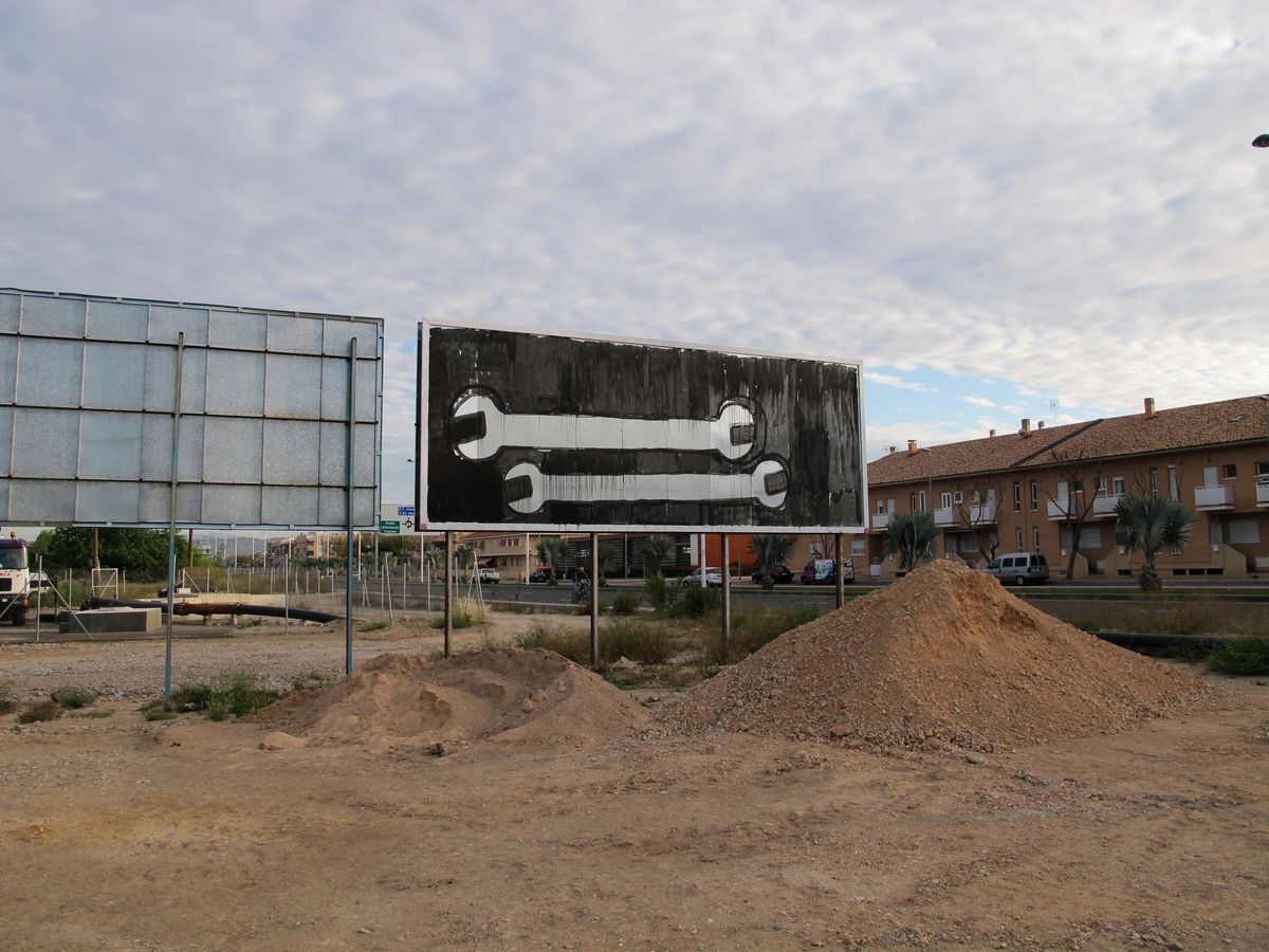 Black and white billboard painted as a large adjustable wrench lying horizontally, black paint dripping down the sign, dirt mounds in front.