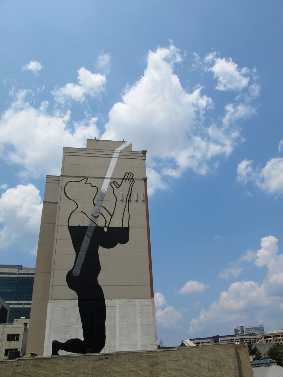 Black outline mural of a kneeling figure with head tilted back and hands raised, a thin white diagonal beam of light painted from the sky down toward the raised hands, on the side of a tall tan building.
