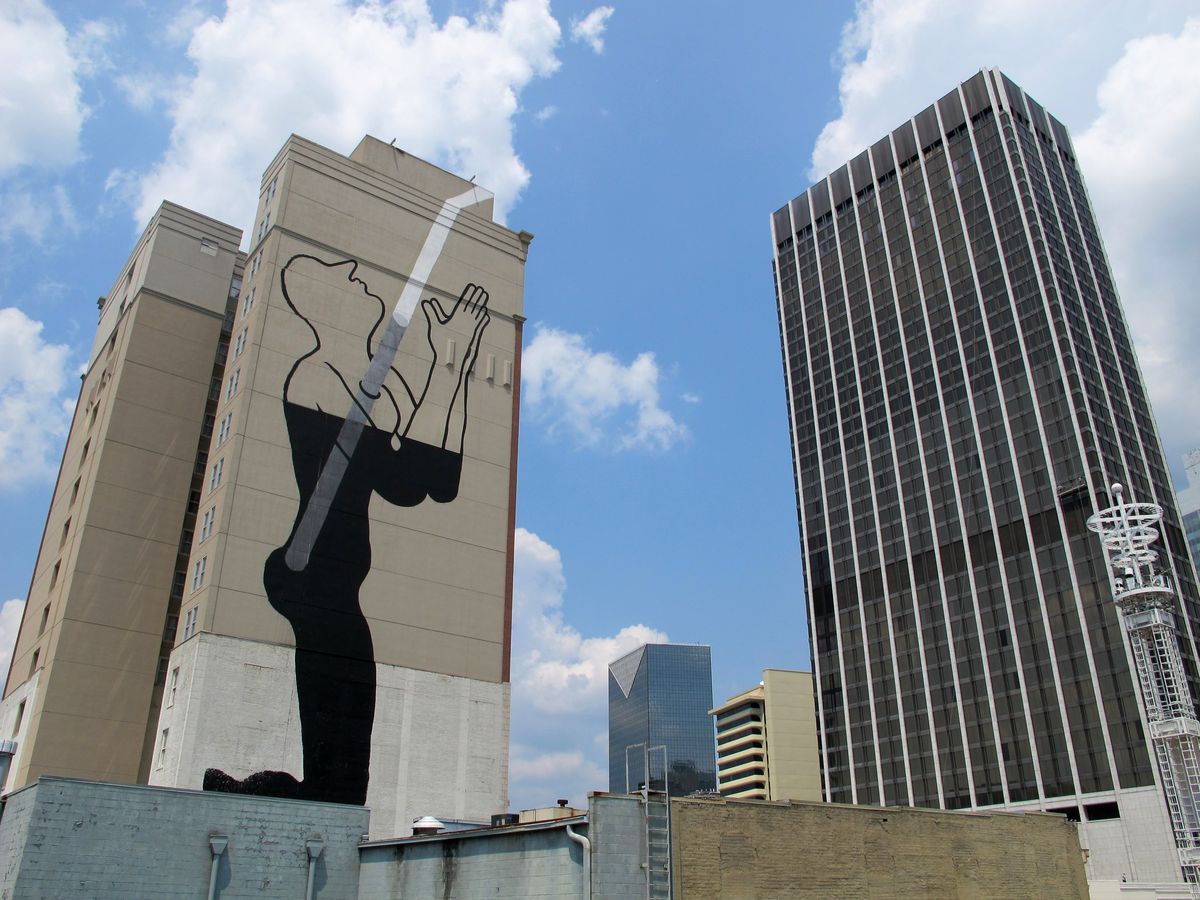 Two tall beige apartment towers, the closer one painted with a black silhouette of a kneeling figure with head tilted back and hands raised to catch a white diagonal beam of light, skyscrapers in the background.