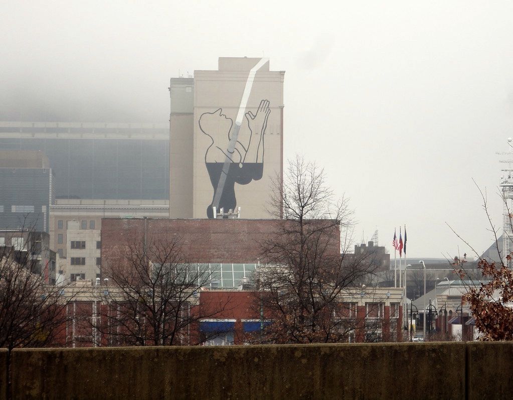 Close-up of the same tower mural in progress, a white diagonal beam painted down the facade toward two open hands, workers on a suspended platform still painting the black silhouette below.